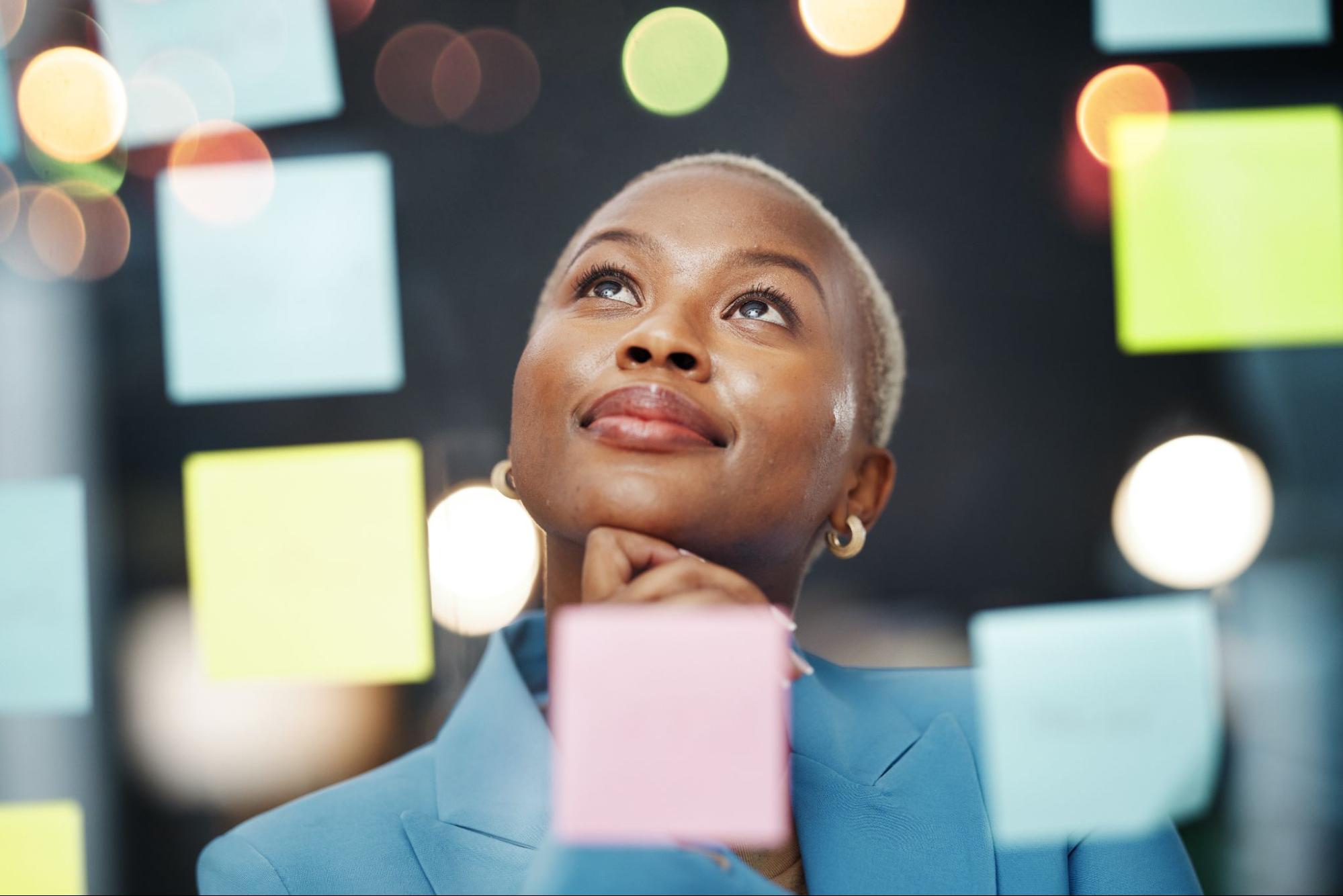 Woman looking up thoughtfully behind colorful sticky notes on a glass board, planning SMART goals for better hormonal health.