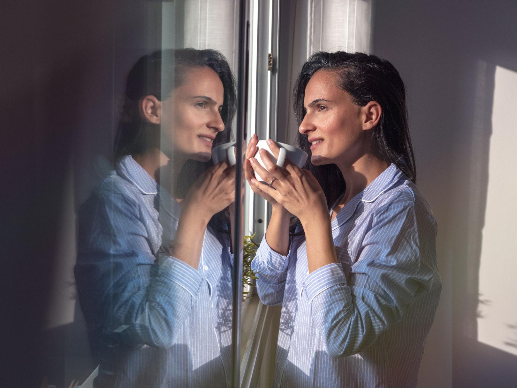 Woman holding a mug by a window, looking at her reflection during a quiet moment of self-reflection.