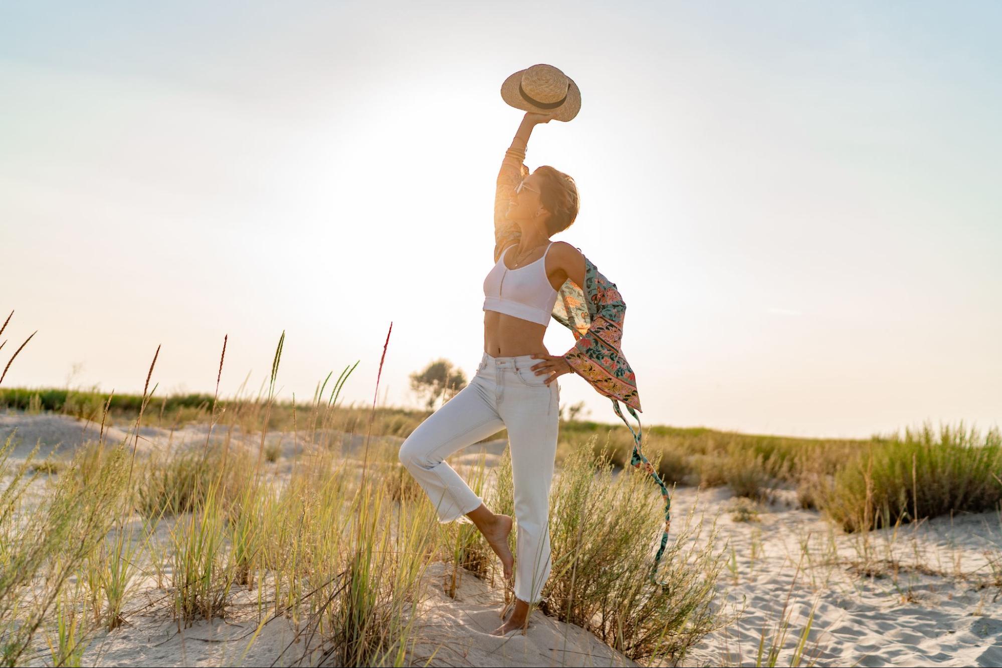 Woman standing on a sandy beach at sunrise with one arm raised holding a hat, symbolizing resilience and nervous system regulation through time outdoors.