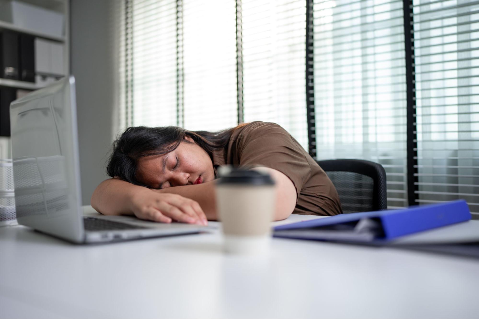 Woman sleeping at her office desk beside an open laptop and a coffee cup, showing daytime fatigue and sleep disruption.
