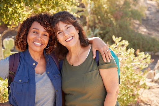 Two middle-aged women smiling with arms around each other during an outdoor hike, wearing backpacks in a sunny nature setting.
