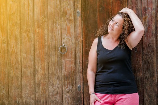 Woman with curly hair standing against a wooden door outdoors, adjusting her hair and looking to the side in warm sunlight.