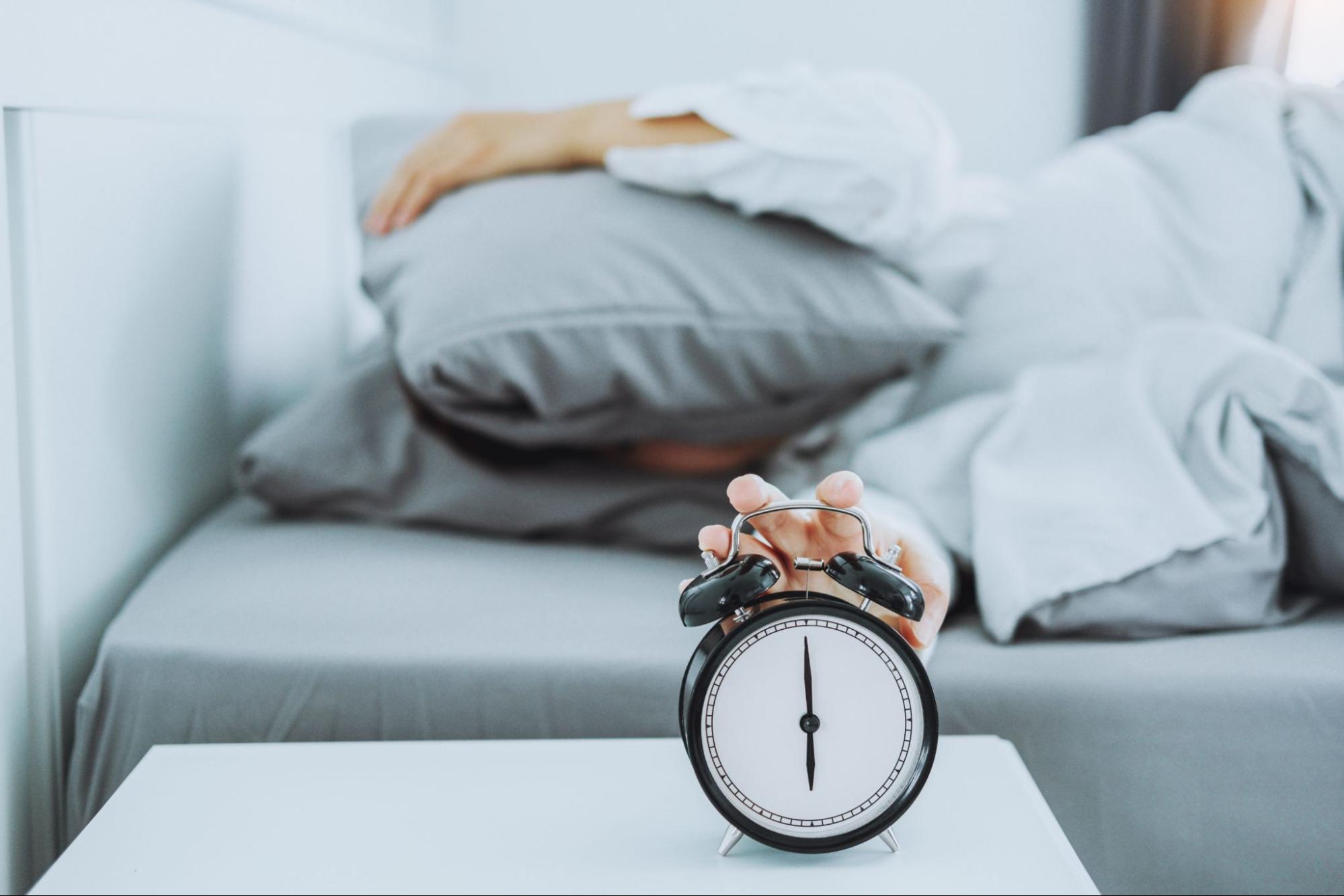 Close-up of an alarm clock with a person’s hand reaching to turn it off, symbolizing the importance of sleep for health and fitness.